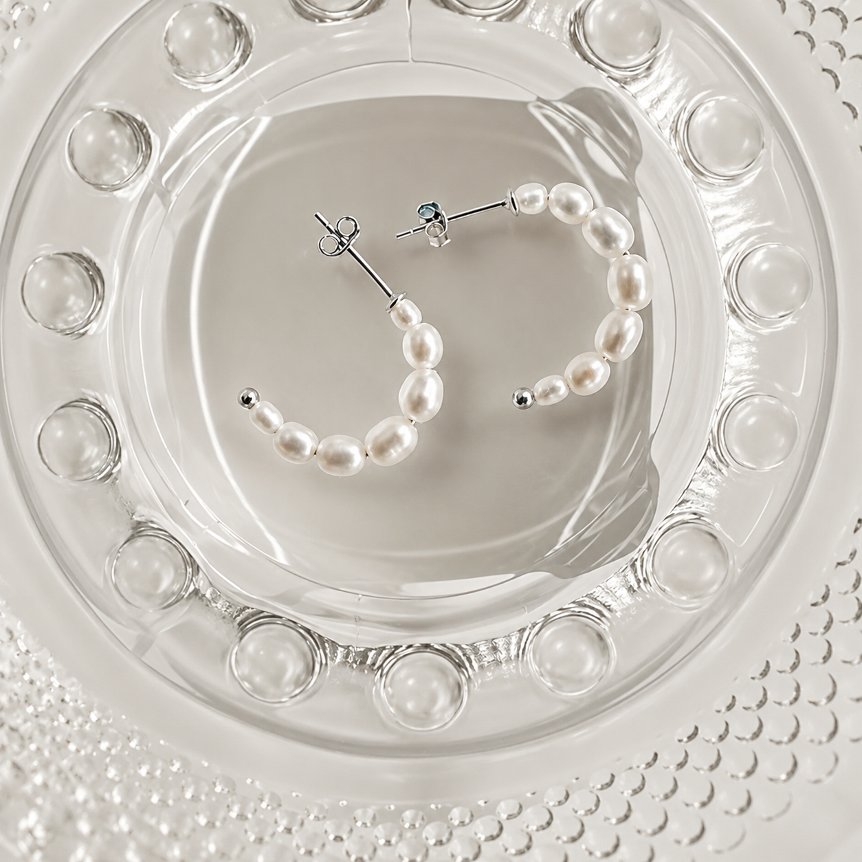 Two silver hoop earrings with white freshwater pearls, displayed on a clear glass plate.